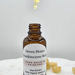 May include: A brown glass bottle with a white label that reads "Green Hojari Frankincense Resin Pure Nourish Skincare Oleo Extract 100% Organic Jojoba Oil External use only". The bottle is sitting on a white surface with small pieces of amber-colored resin.