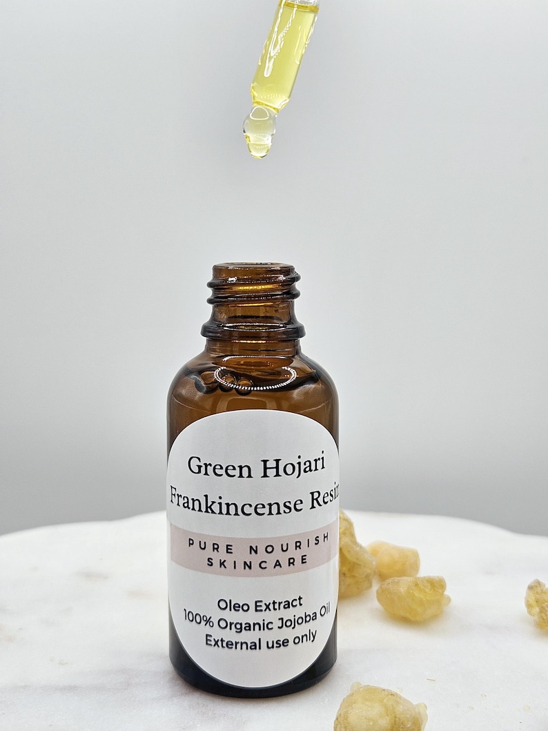 May include: A brown glass bottle with a white label that reads "Green Hojari Frankincense Resin Pure Nourish Skincare Oleo Extract 100% Organic Jojoba Oil External use only". The bottle is sitting on a white surface with small pieces of amber-colored resin.