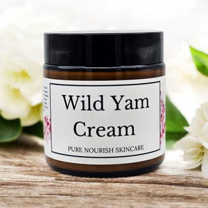 May include: A brown glass jar with a black lid, labelled "Wild Yam Cream" in black text. The label also includes "PURE NOURISH SKINCARE" in smaller text. Pink floral accents are on the label. The jar is on a wooden surface.