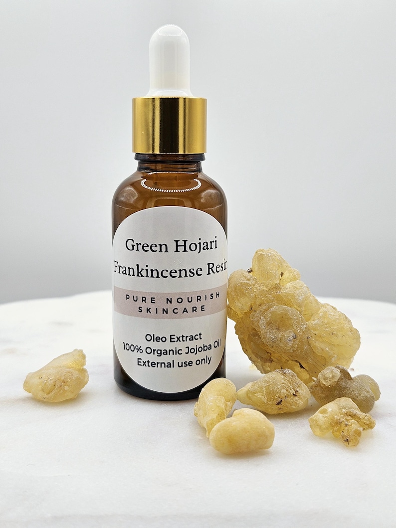 May include: A brown glass bottle with a gold lid and dropper, labeled "Green Hojari Frankincense Resin Pure Nourish Skincare Oleo Extract 100% Organic Jojoba Oil External use only". The bottle is surrounded by small, yellow, resin pieces.