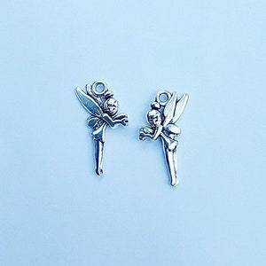Peut inclure: Deux breloques argentées en forme de fée, avec des ailes détaillées et une boucle pour les attacher à des bijoux ou des créations artisanales. Les fées sont en position debout, tenant quelque chose dans leurs mains. Le fond est bleu clair.