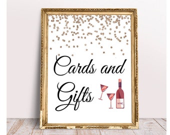 May include: A gold-framed sign with a white background and gold glitter confetti. The sign says "Cards and Gifts" in black cursive font. There are two wine glasses and a bottle of wine with the word "Wine" on the label.