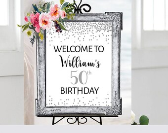 May include: A silver framed welcome sign with a floral design and silver glitter confetti. The sign reads "Welcome to William's 50th Birthday".