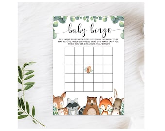 May include: A printable baby bingo card with a woodland theme. The card has a 5x5 grid with spaces for gifts. The card is decorated with a eucalyptus wreath and woodland animals.