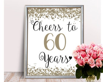 May include: A white sign with a silver frame and gold glitter background. The sign says "Cheers to 60 Years" in black script font. The number 60 is in a gold color.