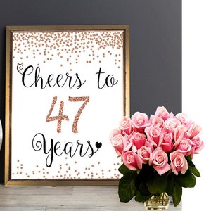 以下が含まれることがあります： 「Cheers to 47 Years」の文字が黒い筆記体とローズゴールドのグリッターで描かれた額入りのプリント。プリントはローズゴールドの紙吹雪と小さな黒いハートで装飾されています。ピンクのバラの花束が額入りのプリントの横に置かれています。