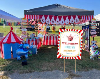 Op de afbeelding: Een feestelijke buitencarnavalscène met een rood-wit gestreepte tent en bewegwijzering. Een bord leest: "Welkom op Olivia's 8e verjaardag." Decoraties zijn onder meer een blauwe olifantenballon, een zebra en een kleine circustent.