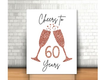 May include: A white print featuring two champagne flutes with rose gold glitter, clinking together. The number "60" and the words "Cheers to Years" are in black script. The print is displayed on a wooden surface against a white brick wall.