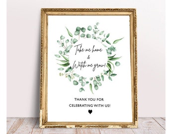 May include: A printable sign with a gold frame and a white background. The sign features a wreath of green eucalyptus leaves and the text "Take me home & Watch me grow!" with a black heart below the text. The text "Thank you for celebrating with us!" is printed below the wreath.