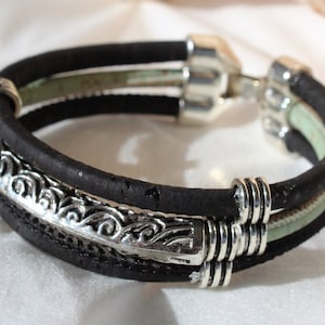May include: A black cork bracelet with a silver decorative band and a silver clasp. The bracelet is made of three layers of cork and has a light green layer underneath the top layer of cork.