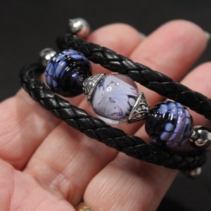 May include: A black leather bracelet with three glass beads. The beads are a swirl of white and purple.