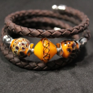 May include: A brown leather bracelet with three colorful glass beads. The beads are orange, yellow, and brown with intricate patterns.