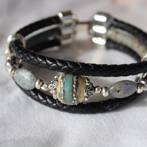 May include: A black leather bracelet with silver accents and a turquoise and white bead. The bracelet has two strands of leather and is adorned with oval-shaped stones.
