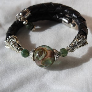 Puede incluir: Un brazalete de cuero negro con detalles de cabeza de dragón plateados y una cuenta de vidrio verde y marrón.