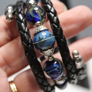 以下が含まれることがあります： 青と白のガラスビーズとシルバーのアクセントが付いた、黒い革のブレスレットです。