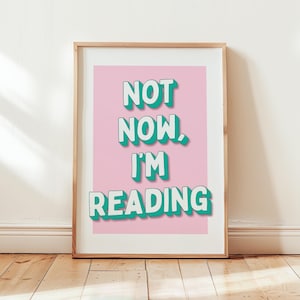 Könnte beinhalten: Ein gerahmter Druck mit rosa Hintergrund und grünem Text, der "NOT NOW, I'M READING" lautet.