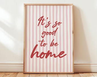 Könnte beinhalten: Ein gerahmter Druck mit einem hellen Holzrahmen. Das Kunstwerk zeigt einen rosa-weiß gestreiften Hintergrund mit dem roten Text "It's so good to be home". Der Druck lehnt an einer weißen Wand auf einem Holzboden.
