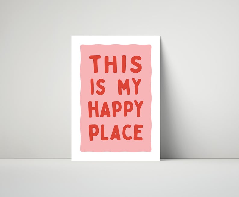 Pu&ograve; includere: Una stampa rettangolare bianca con la scritta "THIS IS MY HAPPY PLACE" in lettere rosse su sfondo rosa. La stampa &egrave; appoggiata a un muro bianco.