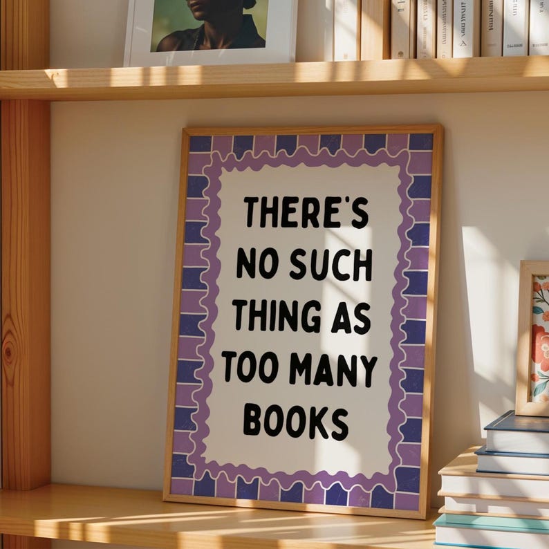 May include: A framed print with the text "THERE'S NO SUCH THING AS TOO MANY BOOKS" in black on a white background. The frame has a decorative purple and blue border. The print is displayed on a wooden shelf.
