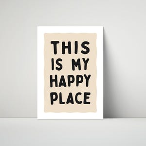 Pu&ograve; includere: Stampa incorniciata con la scritta "THIS IS MY HAPPY PLACE" in grassetto nero. Il testo &egrave; posto su uno sfondo beige chiaro di forma irregolare, con un bordo bianco. La stampa &egrave; esposta su una superficie bianca.