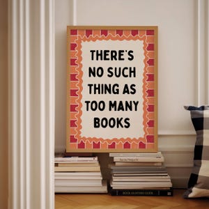 May include: A framed print with the text "THERE'S NO SUCH THING AS TOO MANY BOOKS" in black. The frame is brown with a decorative orange and red border. The print is propped up against a wall, with stacks of books in front.