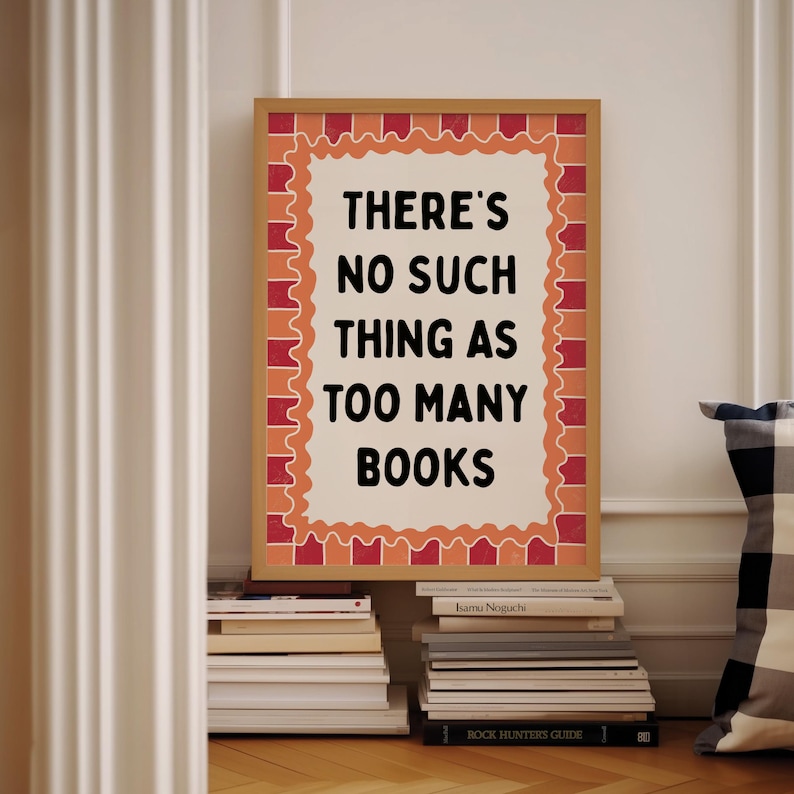 May include: A framed print with the text "THERE'S NO SUCH THING AS TOO MANY BOOKS" in black. The frame is brown with a decorative orange and red border. The print is propped up against a wall, with stacks of books in front.