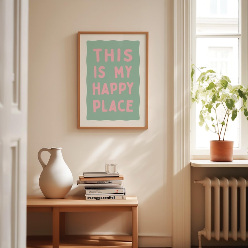 Pu&ograve; includere: Una stampa incorniciata con la scritta "THIS IS MY HAPPY PLACE" in rosa su sfondo verde. La stampa &egrave; in una cornice di legno. Un vaso bianco e una pila di libri sono su un tavolo di legno. La stanza ha una finestra con una pianta.