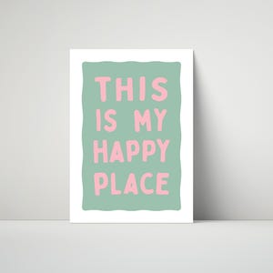 Pu&ograve; includere: Una stampa incorniciata con la scritta "THIS IS MY HAPPY PLACE" in lettere rosa su sfondo verde chiaro. La stampa ha un bordo bianco ed &egrave; esposta contro un muro bianco.