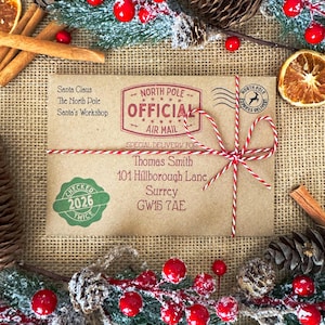Peut inclure: Une enveloppe rectangulaire en papier kraft adress&eacute;e &agrave; Thomas Smith, 101 Hillborough Lane, Surrey, GW15 7AE. Elle porte un tampon rouge North Pole Official Air Mail, un sceau vert Checked Twice 2026 et une ficelle rouge et blanche nou&eacute;e.