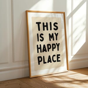 Pu&ograve; includere: Una stampa incorniciata con le parole "THIS IS MY HAPPY PLACE" in grassetto nero. La stampa ha uno sfondo beige e una cornice in legno marrone chiaro. &Egrave; appoggiata a un muro bianco e a un pavimento in legno.