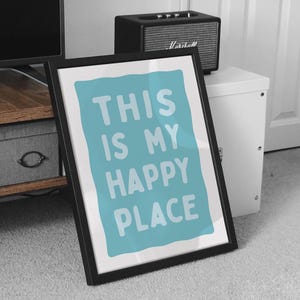 Pu&ograve; includere: Stampa artistica incorniciata con la scritta "THIS IS MY HAPPY PLACE" in lettere bianche su sfondo azzurro. La cornice nera &egrave; angolata contro un tappeto grigio. Un piccolo altoparlante nero &egrave; appoggiato su un mobile bianco.
