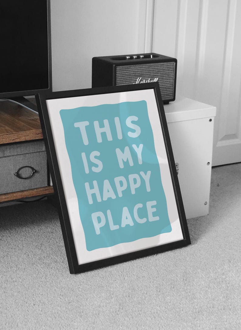Pu&ograve; includere: Stampa artistica incorniciata con la scritta "THIS IS MY HAPPY PLACE" in lettere bianche su sfondo azzurro. La cornice nera &egrave; angolata contro un tappeto grigio. Un piccolo altoparlante nero &egrave; appoggiato su un mobile bianco.