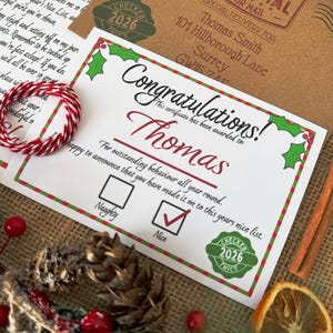 Peut inclure: Un certificat blanc avec une bordure de houx rouge et verte portant le texte Congratulations, Thomas, et une case Nice coch&eacute;e. Le certificat comprend un tampon vert 2026, une enveloppe brune adress&eacute;e &agrave; Thomas Smith et une boucle de ficelle rouge et blanche.