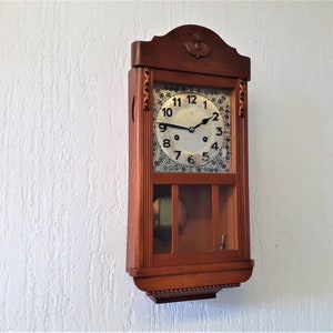 Könnte beinhalten: Eine antike Wanduhr aus Holz mit weißem Zifferblatt und schwarzen römischen Ziffern. Die Uhr hat ein Pendel und eine Glasfront.