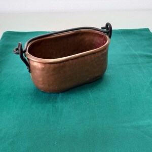 May include: A small copper pot with a hammered finish and a black metal handle. The pot is sitting on a green surface.