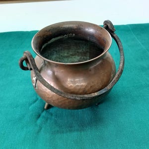 May include: A small, antique copper cauldron with a hammered texture. It has a dark, curved handle and three small feet. The cauldron sits on a teal green cloth.