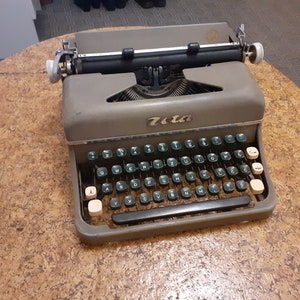 Könnte beinhalten: Eine graue Vintage-Schreibmaschine Zeta mit einem schwarzen Wagenrücklaufhebel und einer grün-weißen Tastatur.