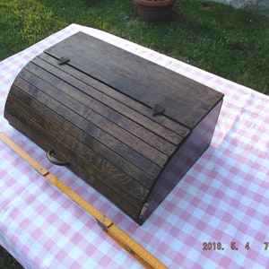 Può includere: Un porta pane in legno vintage con un coperchio incernierato e una maniglia in metallo. La scatola è di colore marrone scuro e ha una parte superiore leggermente curva.