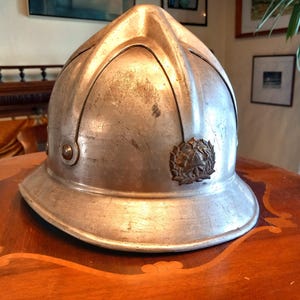 Può includere: Un casco da pompiere vintage color argento con un'ampia tesa e un emblema decorativo. Il casco ha una parte superiore arrotondata con creste verticali. È esposto su un tavolo di legno.