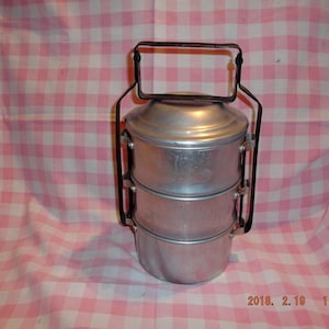 May include: A three-tiered metal lunch box with a black handle. The lunch box is made of silver metal and has a round shape. The lunch box is sitting on a pink and white checkered tablecloth.