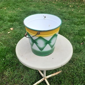 Puede incluir: Un cubo de esmalte blanco con un patr&oacute;n geom&eacute;trico verde y amarillo. El cubo tiene un asa de madera y est&aacute; sentado en una mesa blanca.
