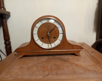 May include: A wooden mantel clock with a white face and gold-coloured hands. The clock has a simple design with a rounded top and a rectangular base.