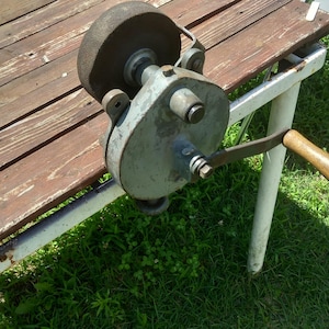 Peut inclure: Une meule à manivelle antique, avec une grande pierre circulaire et un boîtier en métal moulé. La meule est montée sur un banc en bois avec une surface marron et des pieds blancs. Une poignée en bois est attachée.