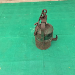 Peut inclure: Une lanterne en métal vert avec une poignée et un bec. La lanterne est rouillée et a une finition usée.