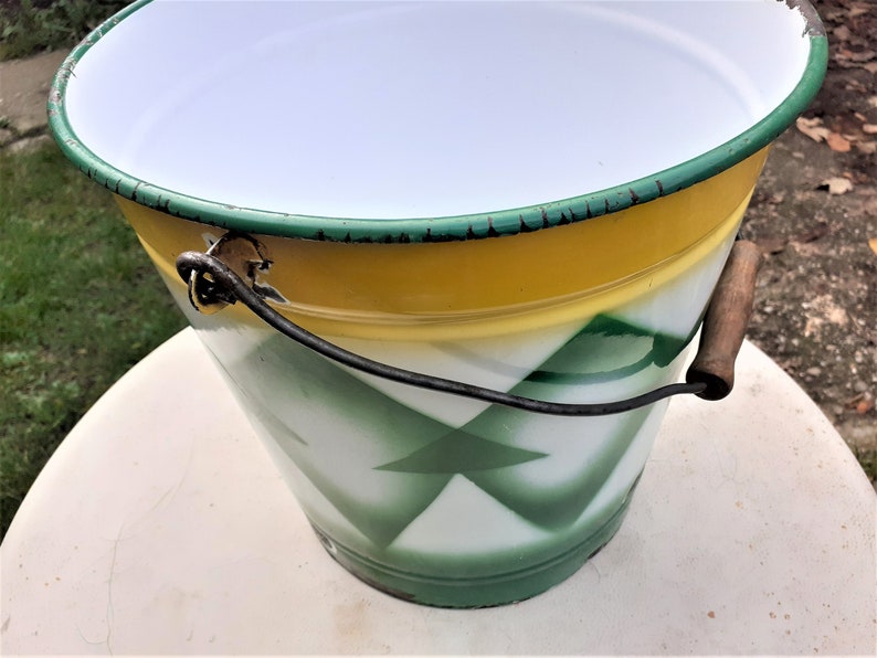 Puede incluir: Un cubo de esmalte vintage con un patr&oacute;n geom&eacute;trico verde, amarillo y blanco. El cubo tiene un asa de metal negro y un asa de transporte de madera.