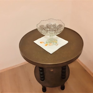 Könnte beinhalten: Ein kleiner, runder Messingtisch mit einer einzigen Ebene und drei Beinen. Eine klare Glasschale mit einem dekorativen Muster steht auf einer weißen Serviette auf dem Tisch.