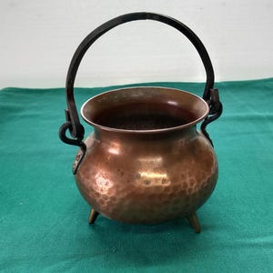 May include: A small copper pot with a hammered finish and a black metal handle. The pot has three short legs and is sitting on a green surface.