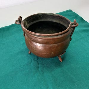 Può includere: Una pentola di rame con tre gambe e due manici. La pentola è su una tovaglia verde.