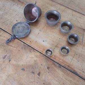 May include: A collection of antique metal measuring cups and a hinged container with a handle, displayed on a wooden surface. The cups are of various sizes, with a tarnished finish. The container is open, showing its interior.