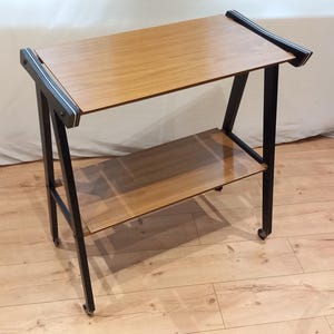 Könnte beinhalten: Ein zweistöckiger Servierwagen mit hellem Holzmaserungsfinish. Der Wagen hat einen schwarzen Metallrahmen und kleine Räder. Die oberen und unteren Regale sind rechteckig. Der Wagen ist ca. 76 cm lang, 46 cm breit und 71 cm hoch.